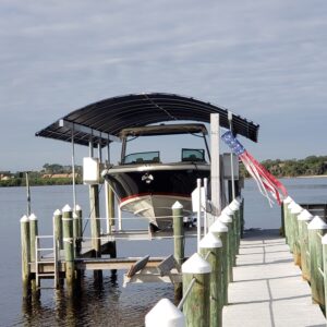 Boat lift canopy protecting a large black boat docked on a sunny Florida waterfront, showcasing the sturdy frame and quality fabric essential for storm season durability.